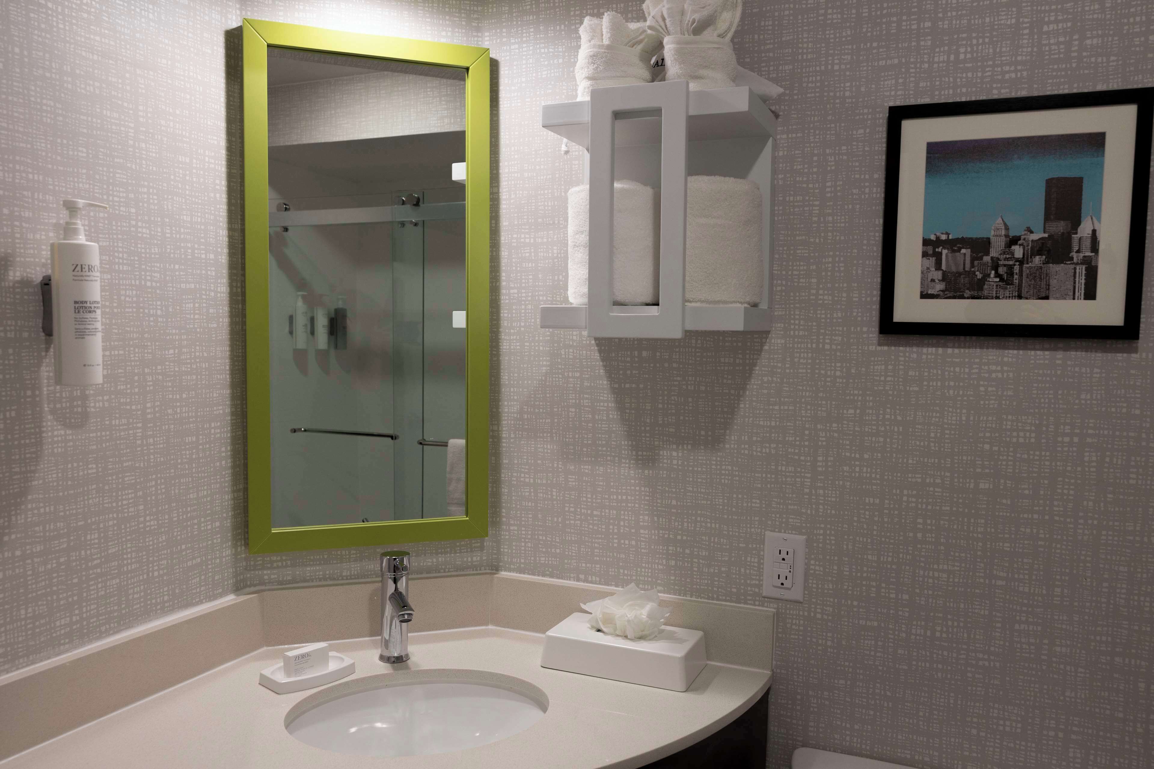 A bathroom with a white sink, a green-framed mirror, a white towel dispenser, and a framed cityscape picture on the wall.