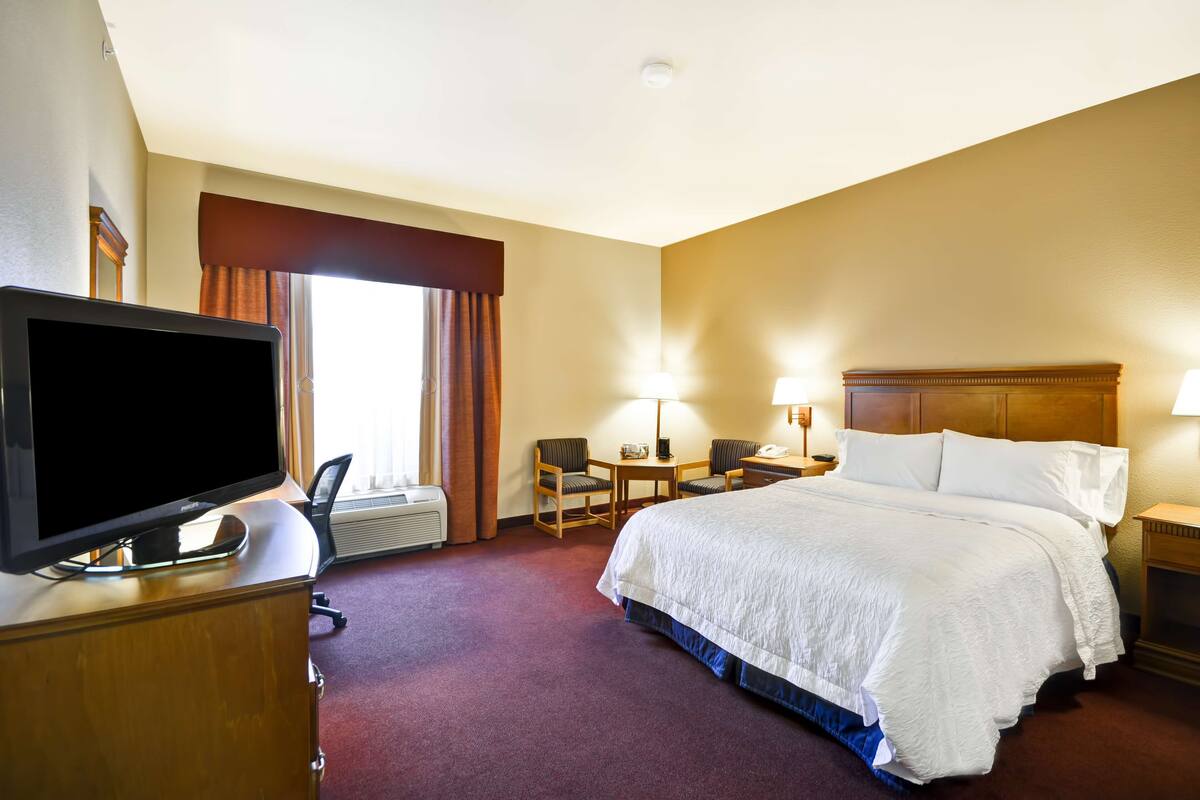 A hotel room with a bed, a desk with a computer, a TV, and a window with curtains.