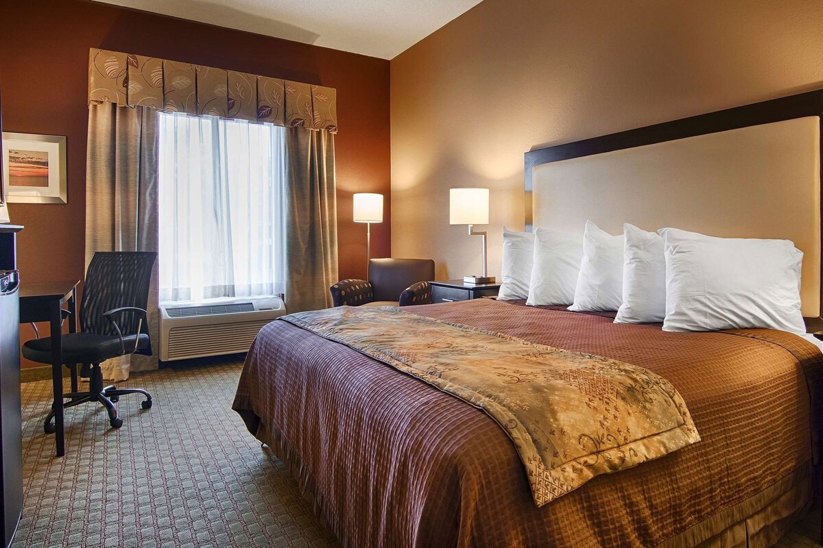 A hotel room with a bed, a desk, a chair, a window with curtains, and a wall-mounted lamp.