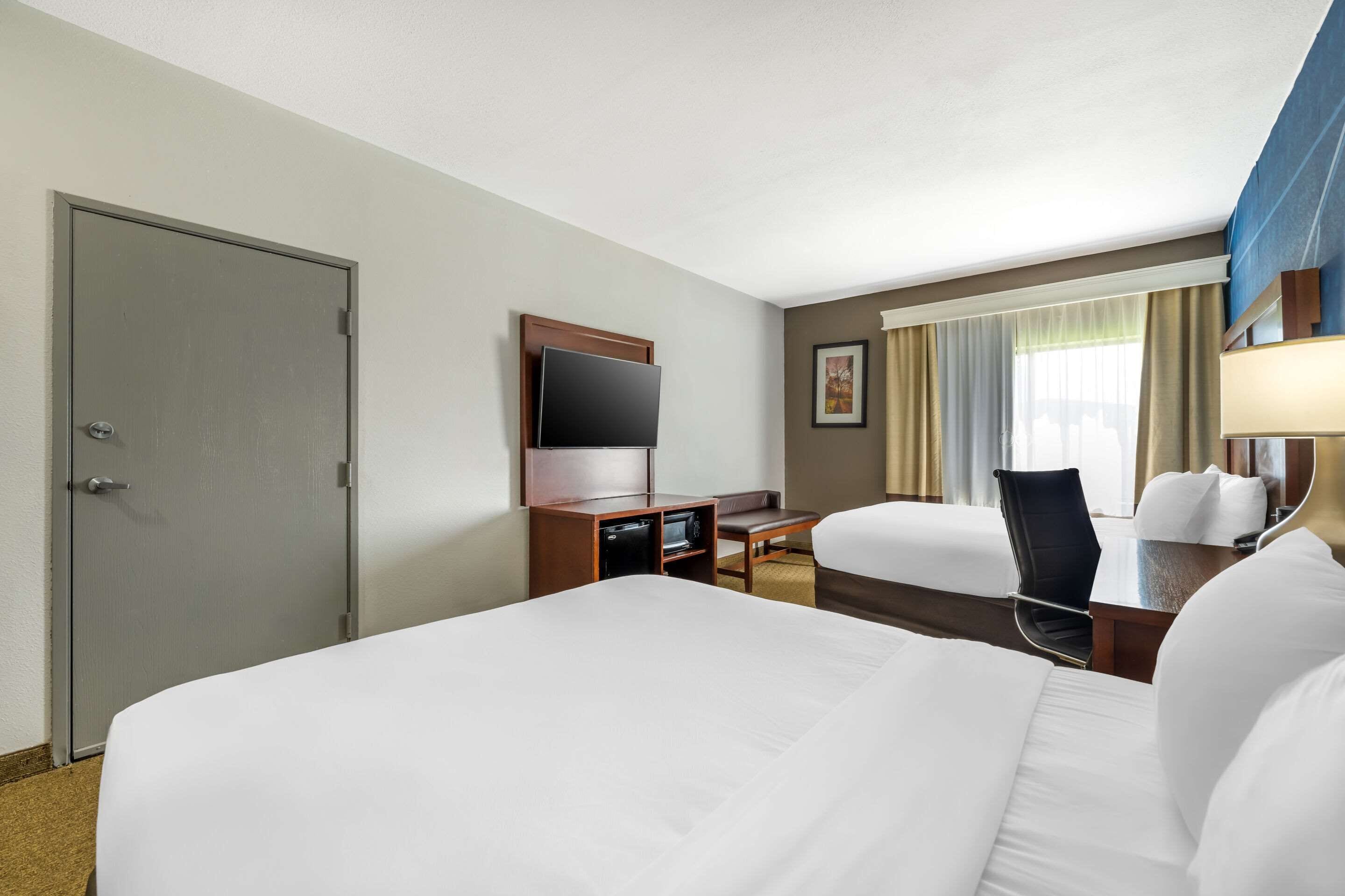 A hotel room with two beds, a desk with a flat-screen TV, a minibar, and a window with curtains.