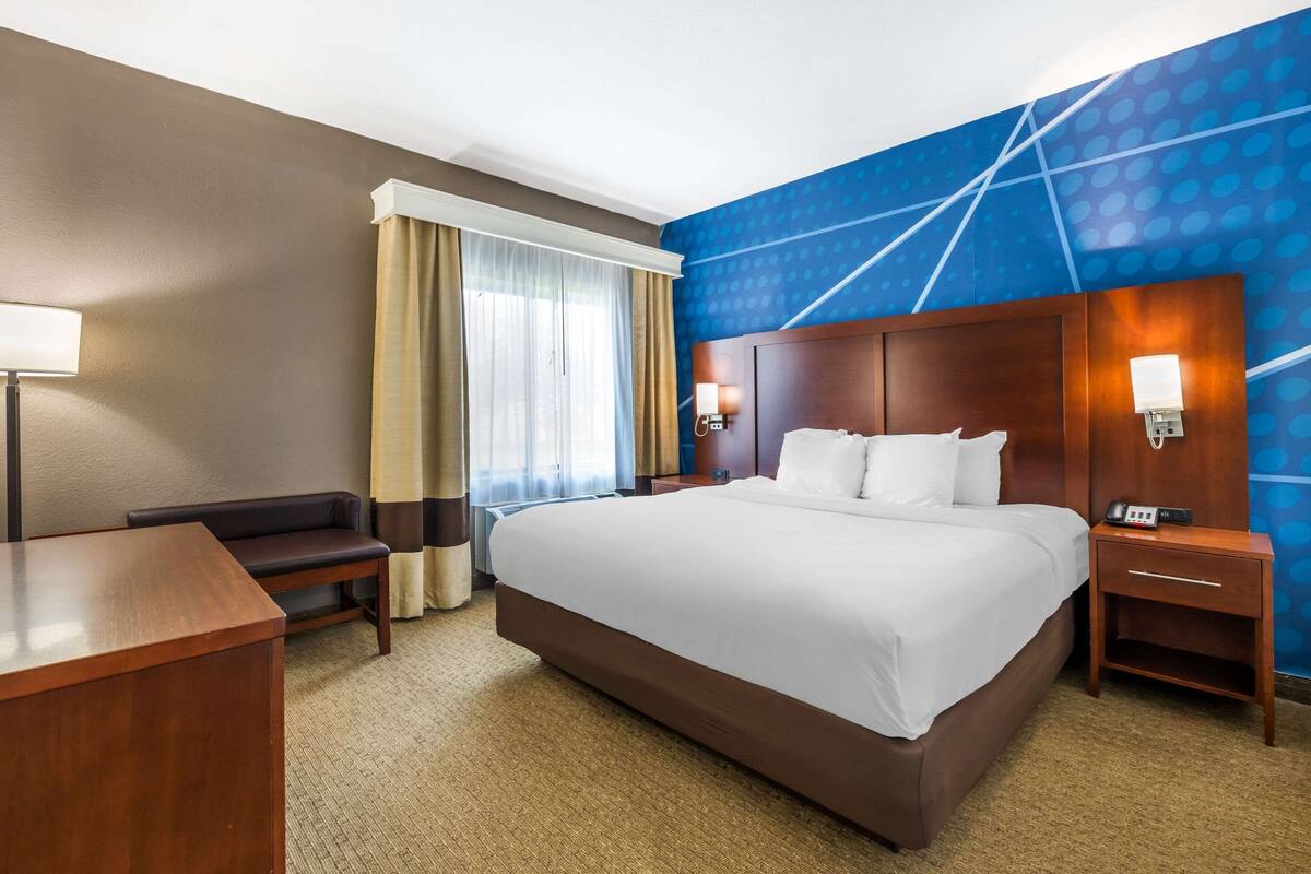 Ein Hotelzimmer mit einem großen Bett, einem Schreibtisch, einem Stuhl, einem Nachttisch mit Lampe und einer blauen Wand mit Muster.