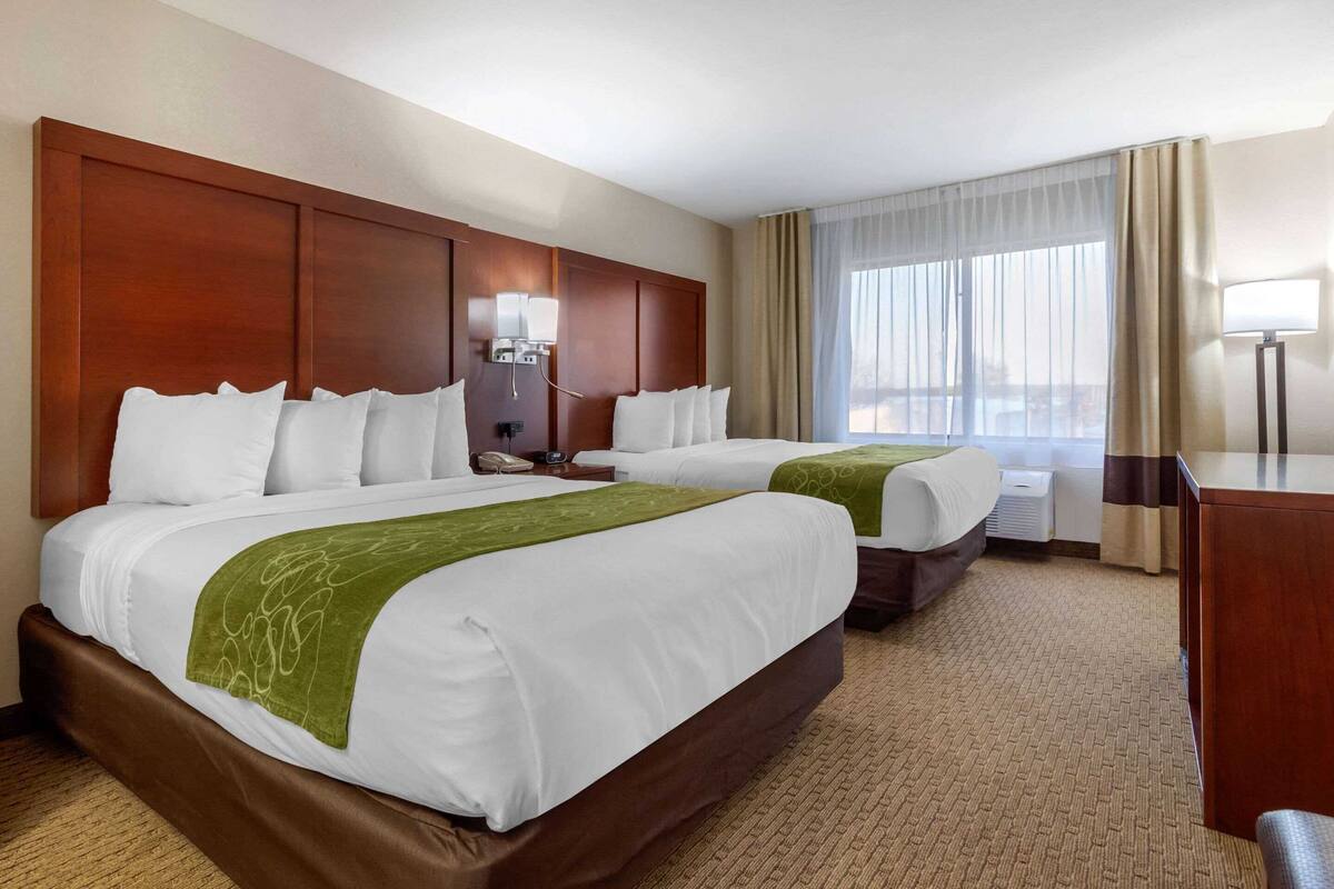 Ein Hotelzimmer mit zwei Betten, einem Schreibtisch, einer Lampe und einem großen Fenster mit Vorhängen.