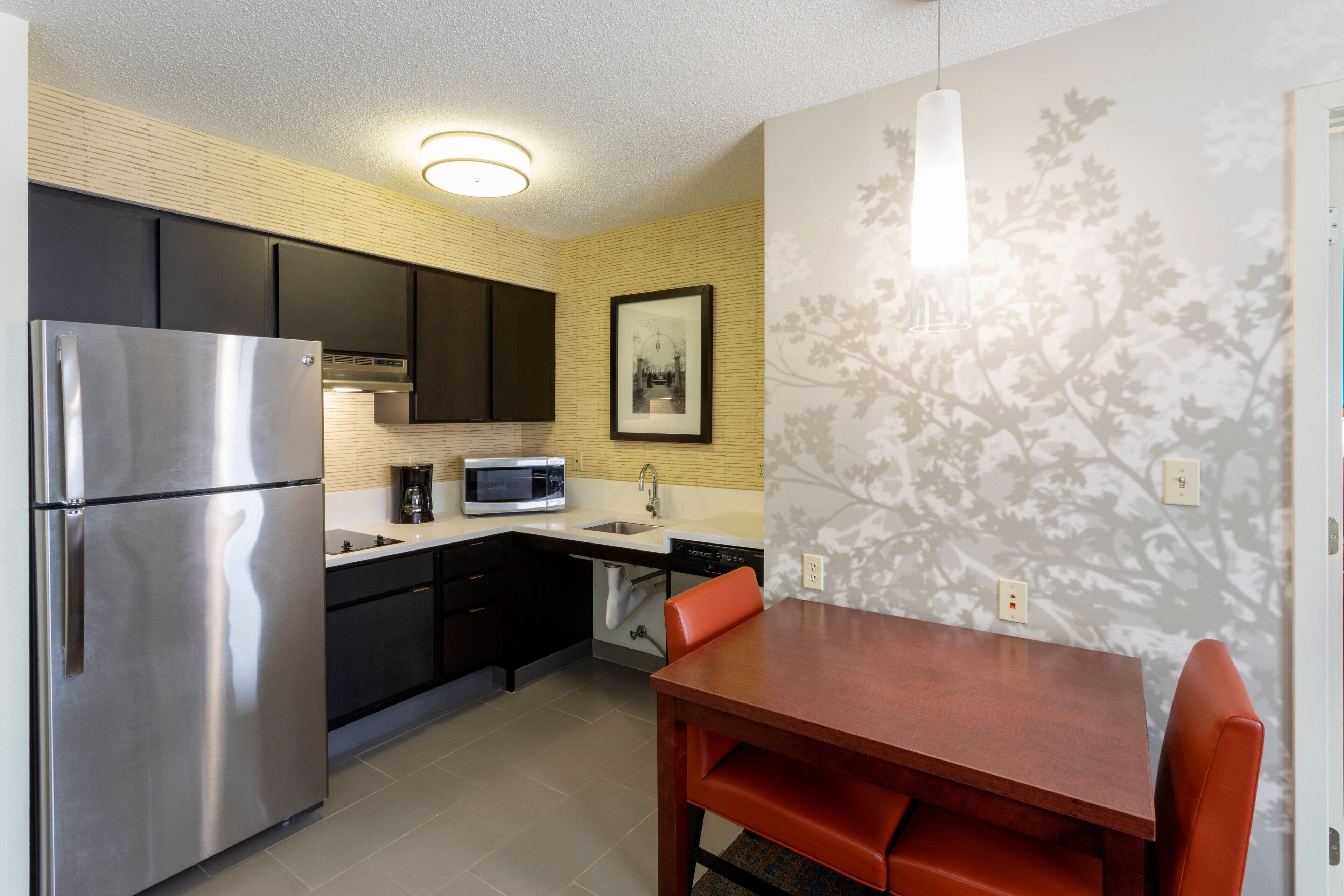 A compact kitchen with a stainless steel refrigerator, a microwave, and a dishwasher. There is a dining table with orange chairs and a wall-mounted light fixture.