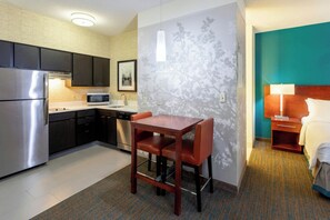 A compact hotel room with a kitchenette, a dining area with a table and chairs, a bed with a nightstand, and a wall-mounted lamp. - Residence Inn by Marriott Minneapolis St. Paul/Roseville (Roseville)