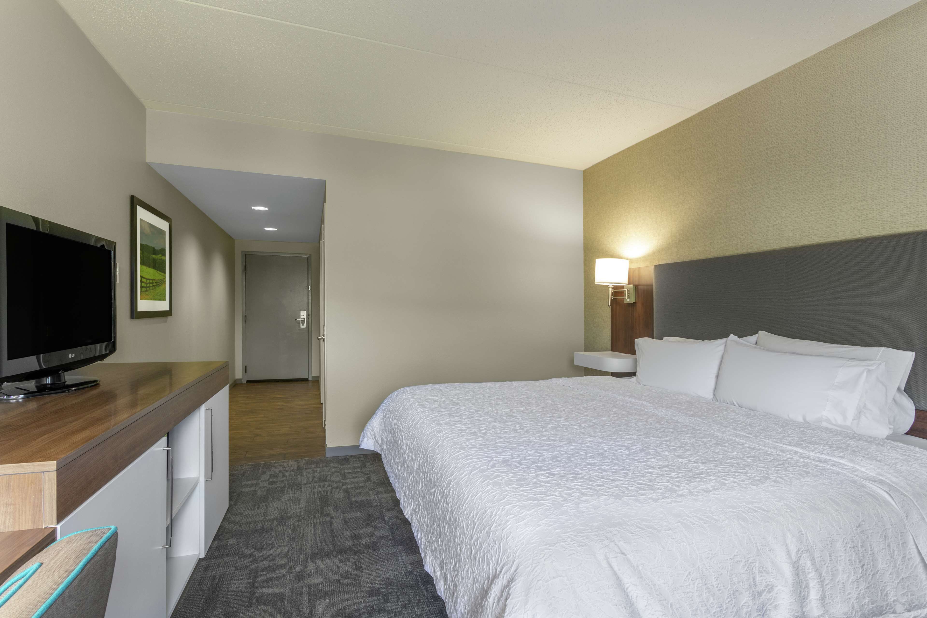 Quarto de hotel com uma cama, uma televisão sobre uma escrivaninha, uma luminária e uma porta que dá para outro quarto.