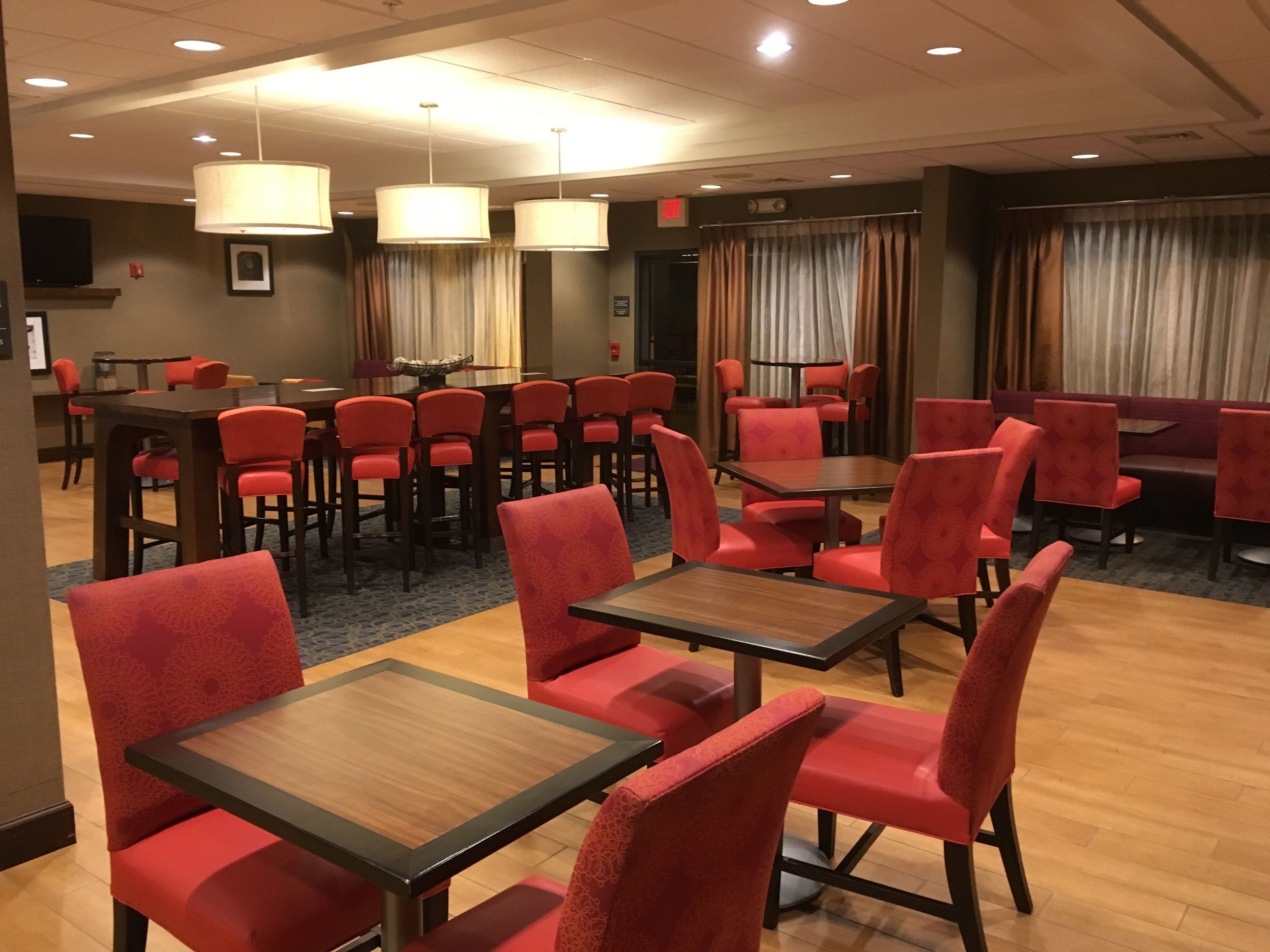 A hotel dining area with red upholstered chairs and tables, a large dining table, and pendant lighting.