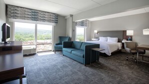 Ein Hotelzimmer mit einem großen Bett, einer Couch, einem Schreibtisch und einem Fernseher. Große Fenster bieten einen Ausblick ins Freie.