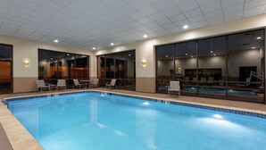 Ein Hallenbad mit klarem Wasser, umgeben von Glaswänden und -türen, ausgestattet mit mehreren Liegestühlen und Tischen.