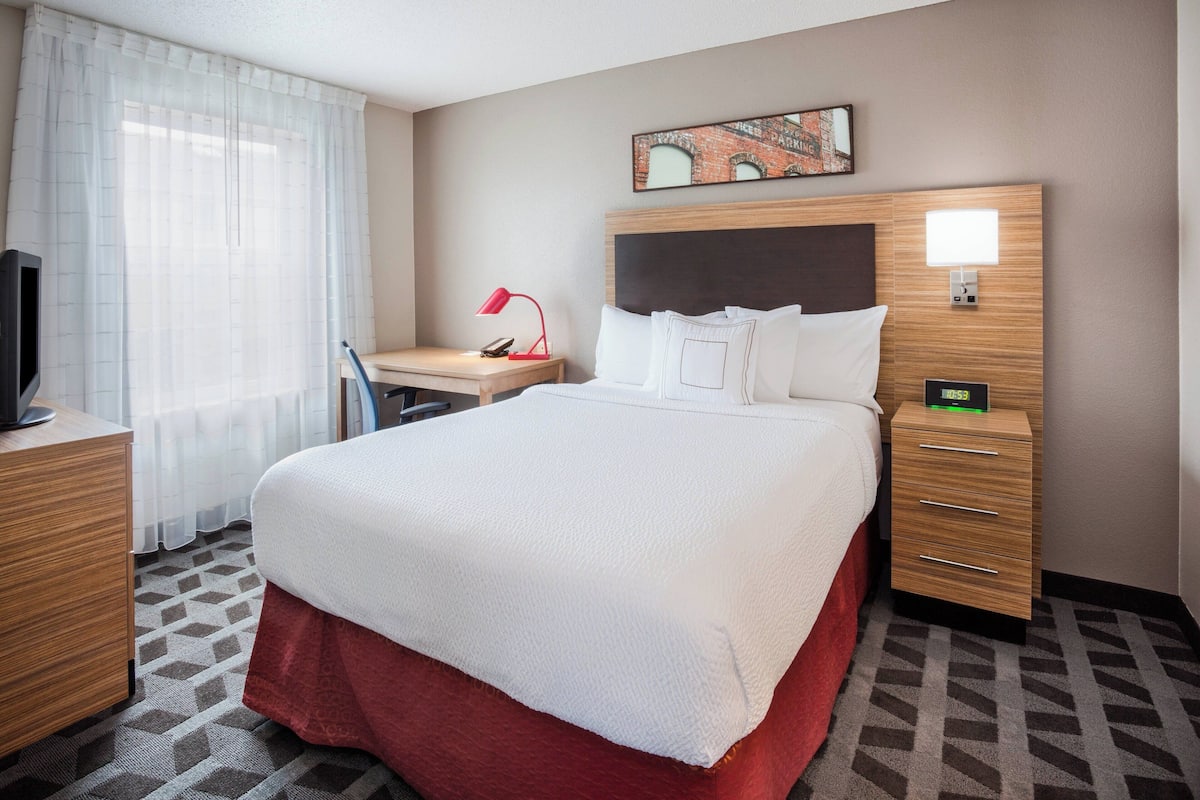 A hotel room with a bed, desk, chair, TV, and a painting on the wall.