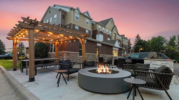 Un patio al aire libre con un hogar circular, asientos y una pérgola con luces colgantes.