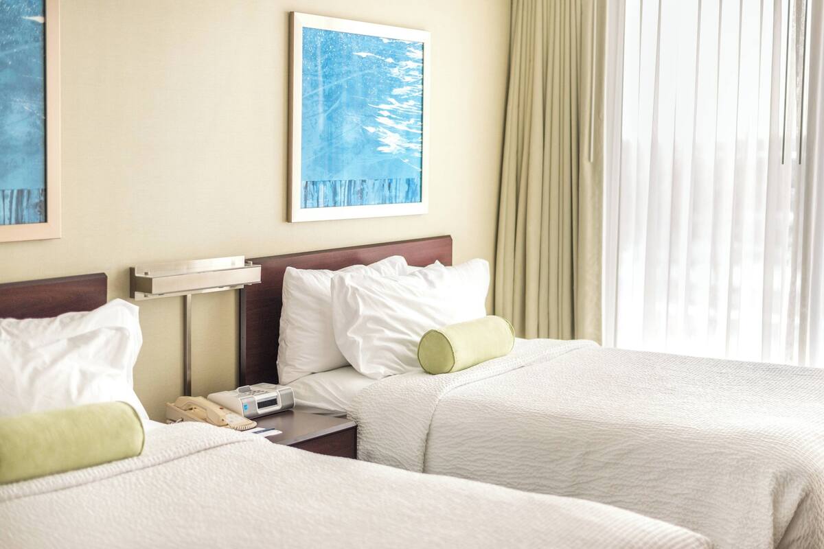 A hotel room with two beds, a bedside table with a phone, a painting on the wall, and a window with curtains.
