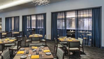 A hotel dining area with tables set for guests, large windows with curtains, and a modern chandelier.