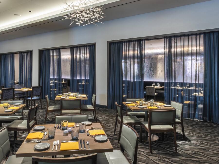 A hotel dining area with tables set for guests, large windows with curtains, and a modern chandelier.