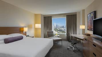 A hotel room with a large bed, a desk, a chair, a TV, and a view of the cityscape.