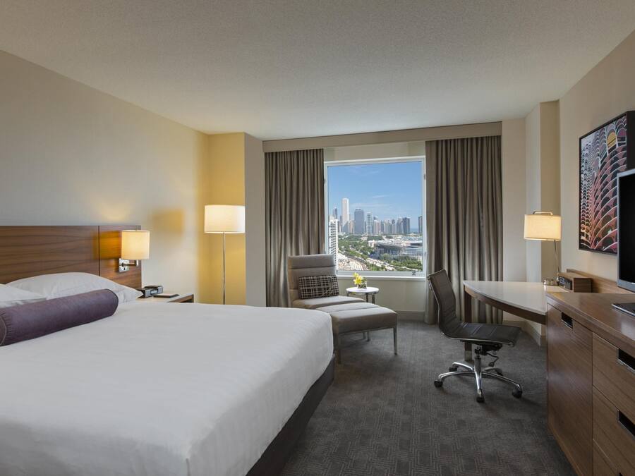 A hotel room with a large bed, a desk, a chair, a TV, and a view of the cityscape.