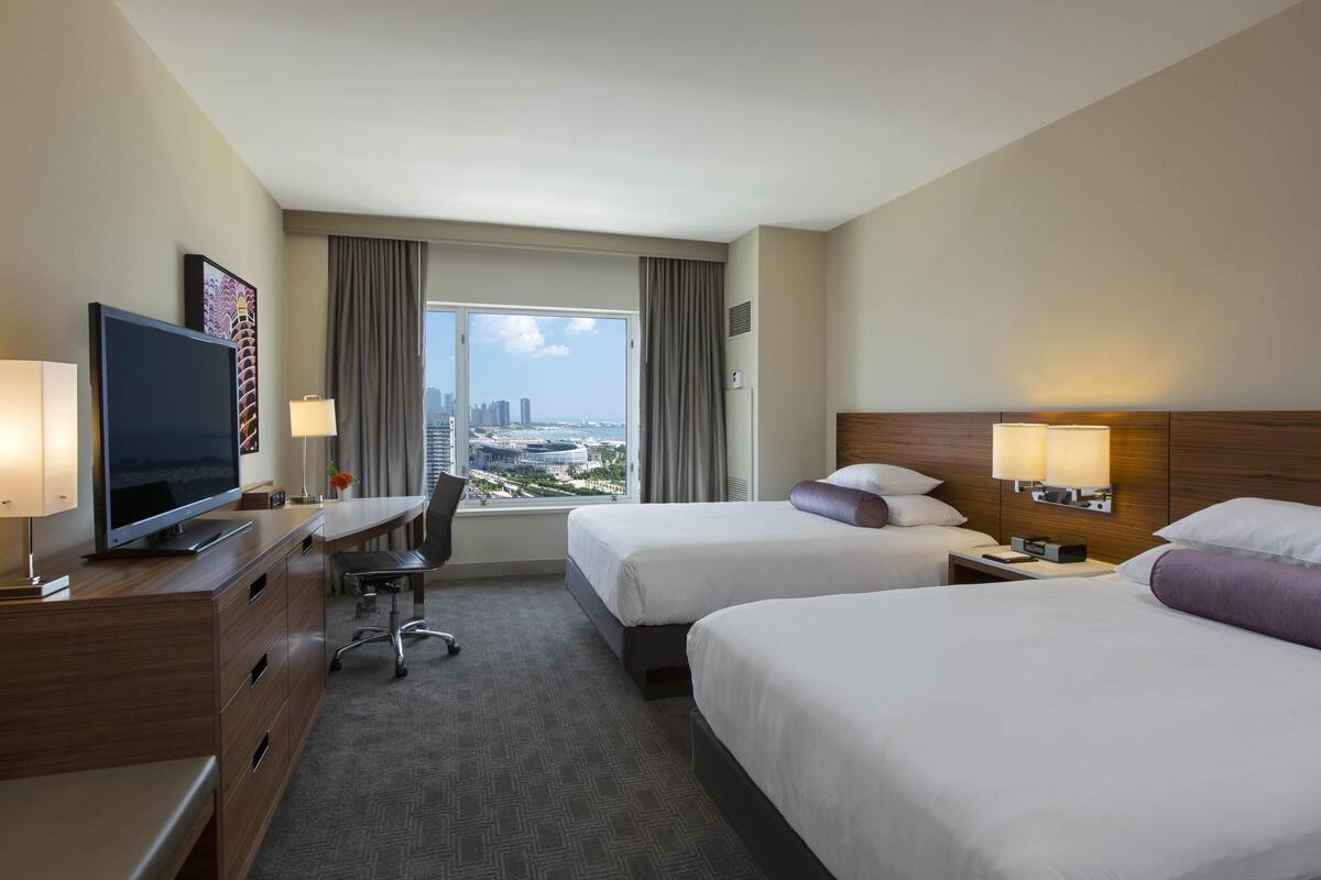 Quarto de hotel com duas camas, uma escrivaninha, televisão e vista para a cidade.