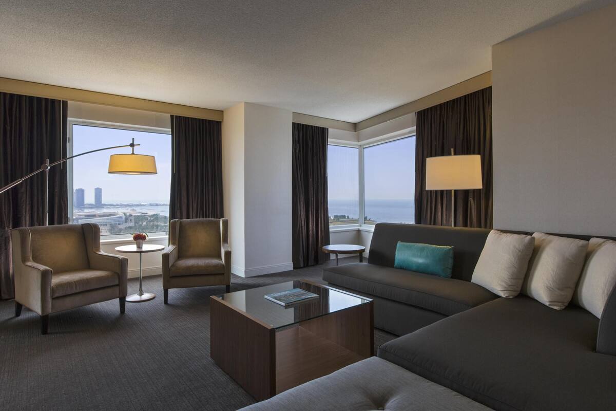 Quarto de hotel com sofá, poltronas, mesa de centro, luminária e vista para a paisagem urbana.