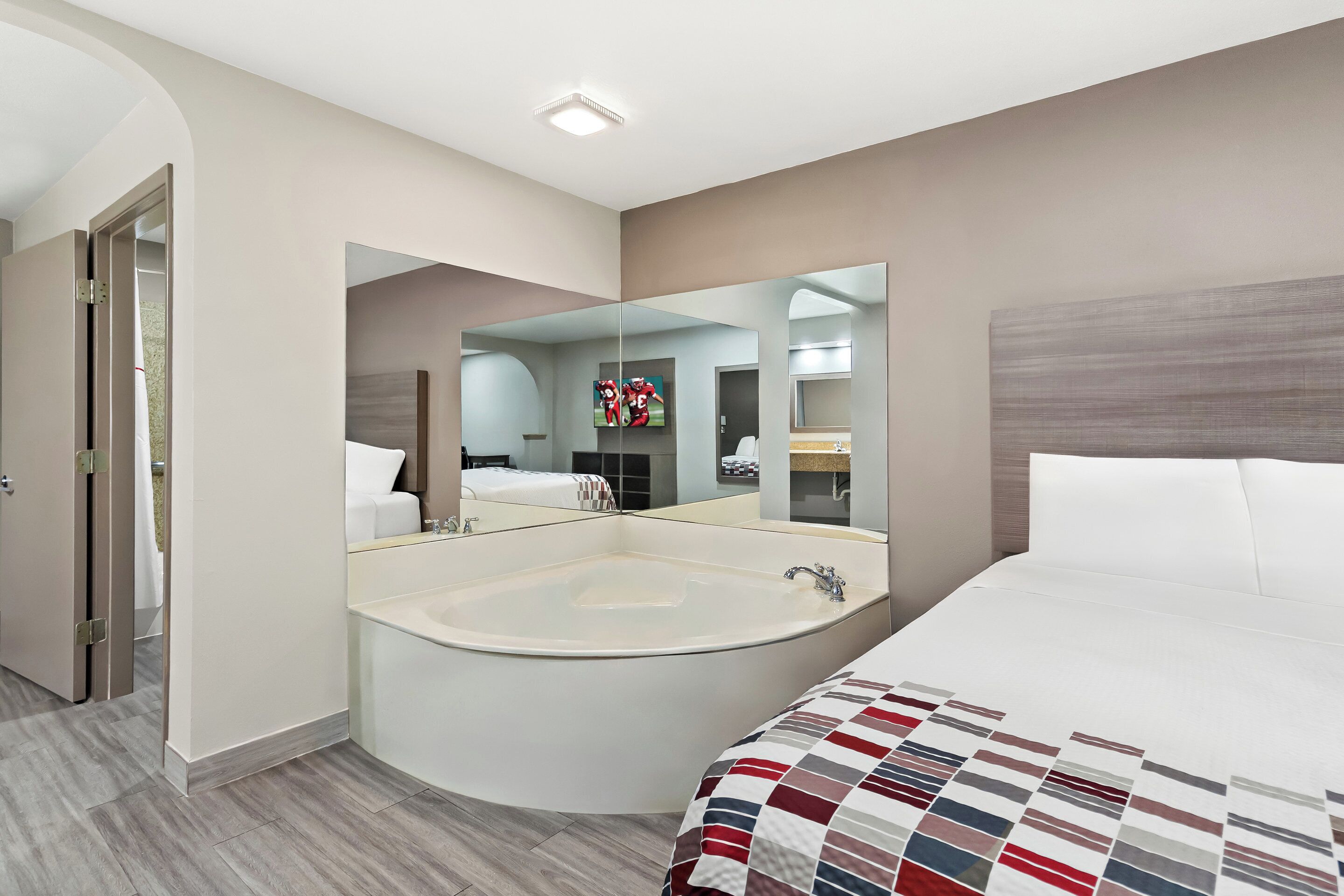 Quarto de hotel moderno com uma cama grande, uma banheira de hidromassagem no canto e um espelho grande que reflete o aposento.