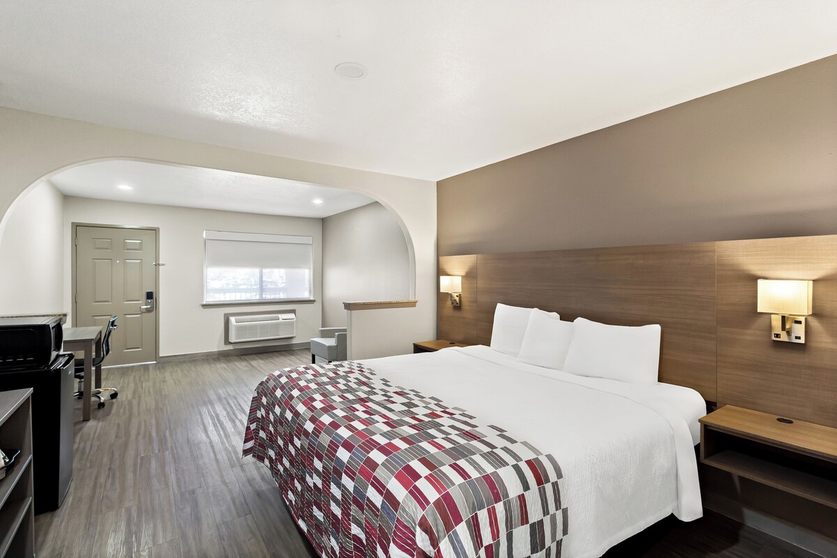 Et hotelværelse med en stor seng, et skrivebord og en stol. Der er et vindue med persienner, en dør og en lampe på væggen.
