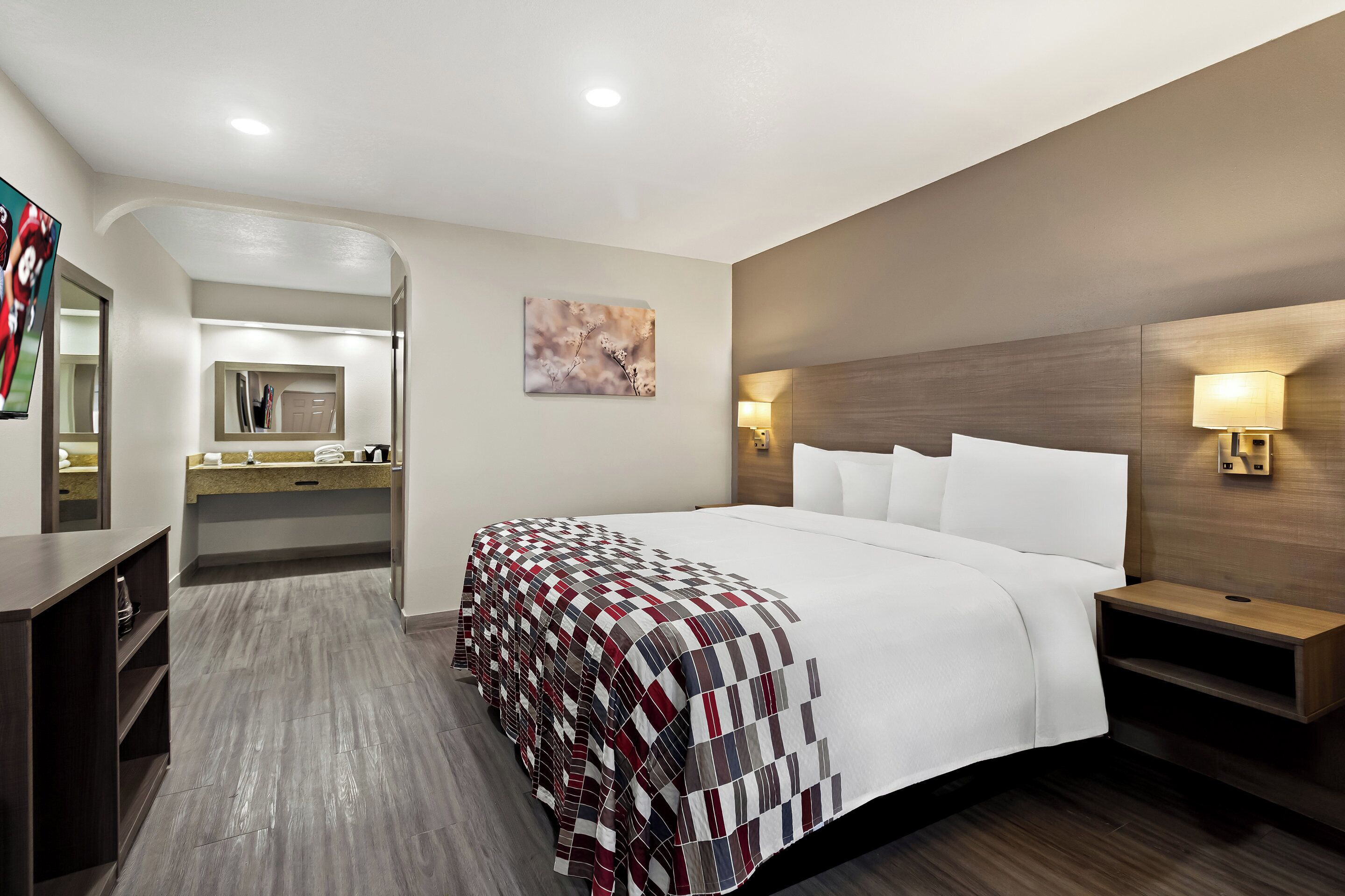 A hotel room with a large bed, a bedside table, a nightstand, a mirror, a vanity with a sink, and a wall-mounted TV.