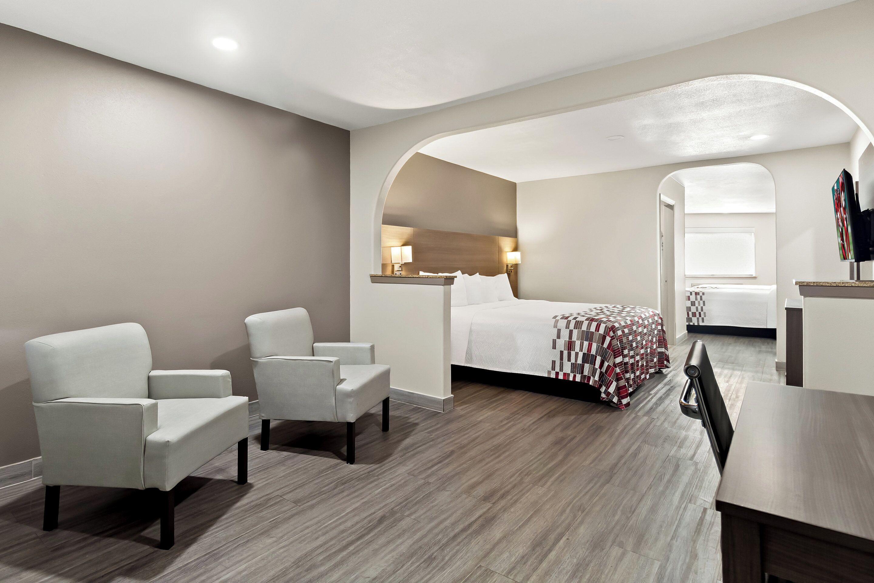 A modern hotel room with a bed, two white armchairs, a wooden desk, and a large window.