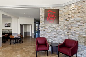 Un hall d’entrée de Red Roof Inn avec un mur en pierre, deux fauteuils rouges et un coin repas avec des tables et des chaises en bois.
