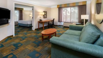 A hotel room with a green sofa, a wooden coffee table, a desk with a chair, a television, and a bed with a blue headboard.