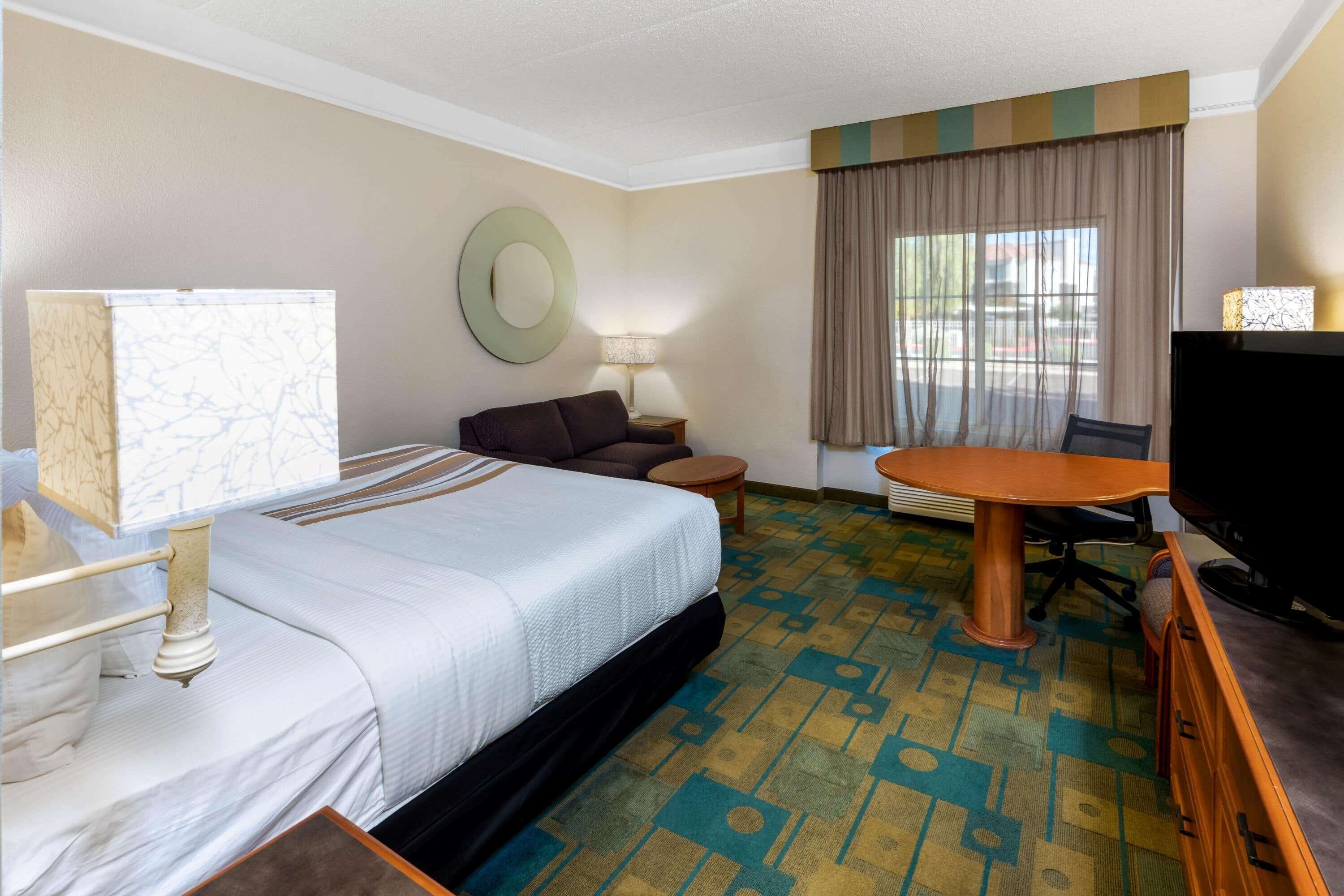Habitación de hotel con cama, escritorio, silla, televisor y ventana con cortinas.