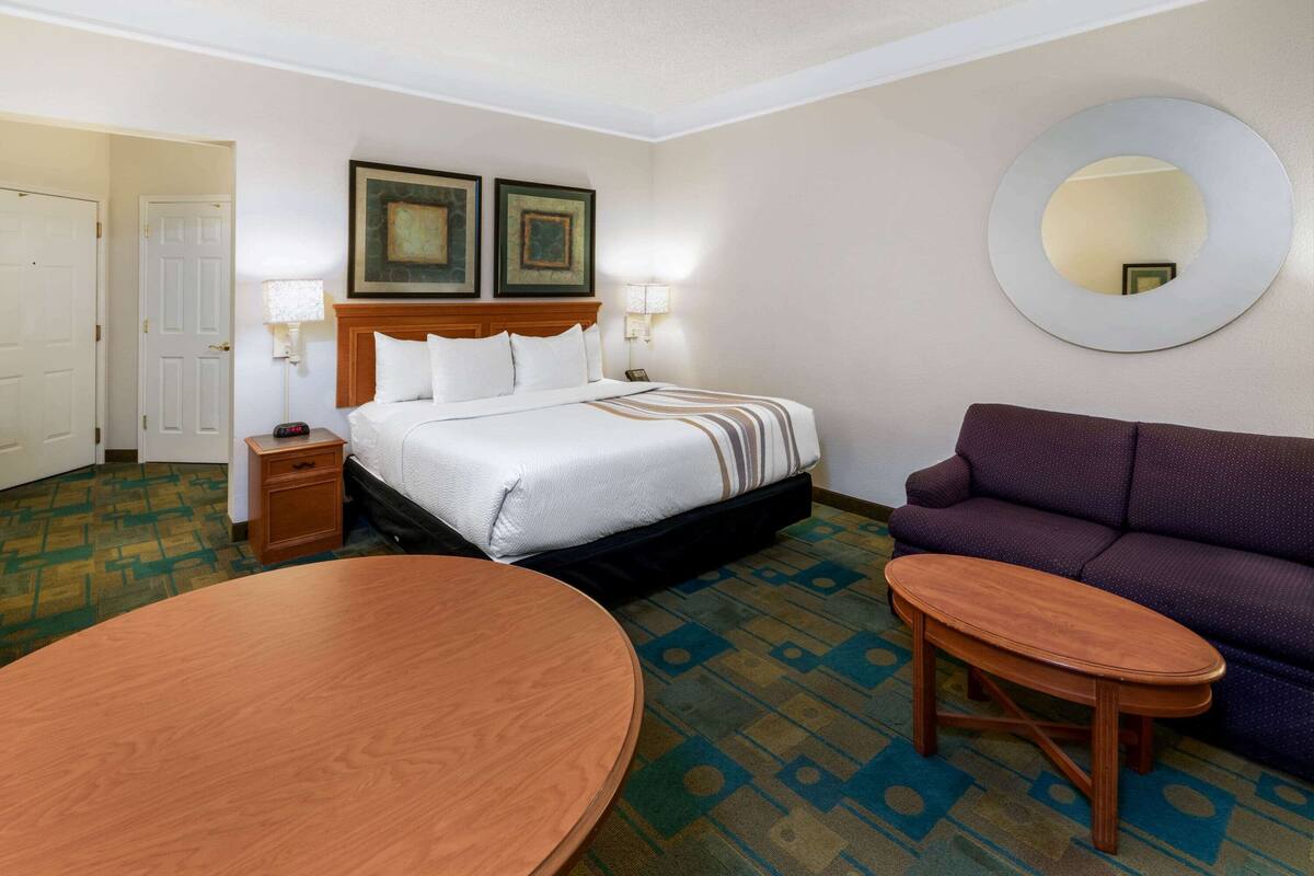 Une chambre d’hôtel avec un lit, une table ronde en bois, un canapé violet et un miroir fixé au mur.