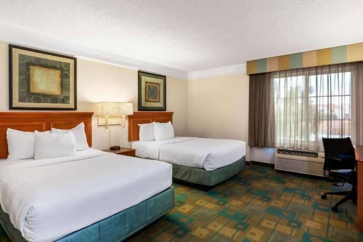 A hotel room with two beds, a desk, a chair, an air conditioning unit, and two framed pictures on the wall.