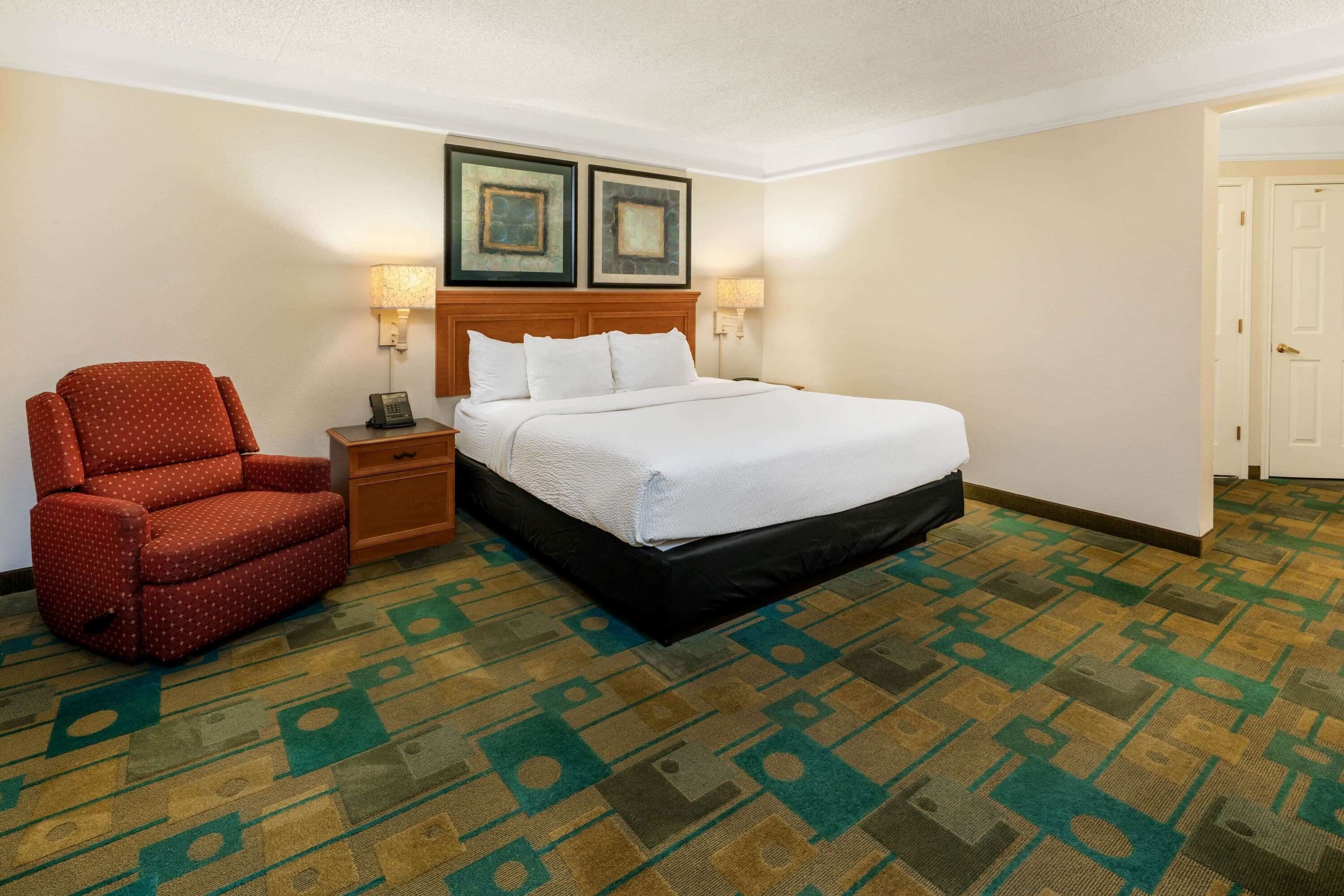 A hotel room with a bed, a red polka dot chair, a nightstand, two wall-mounted lamps, and a patterned carpet.
