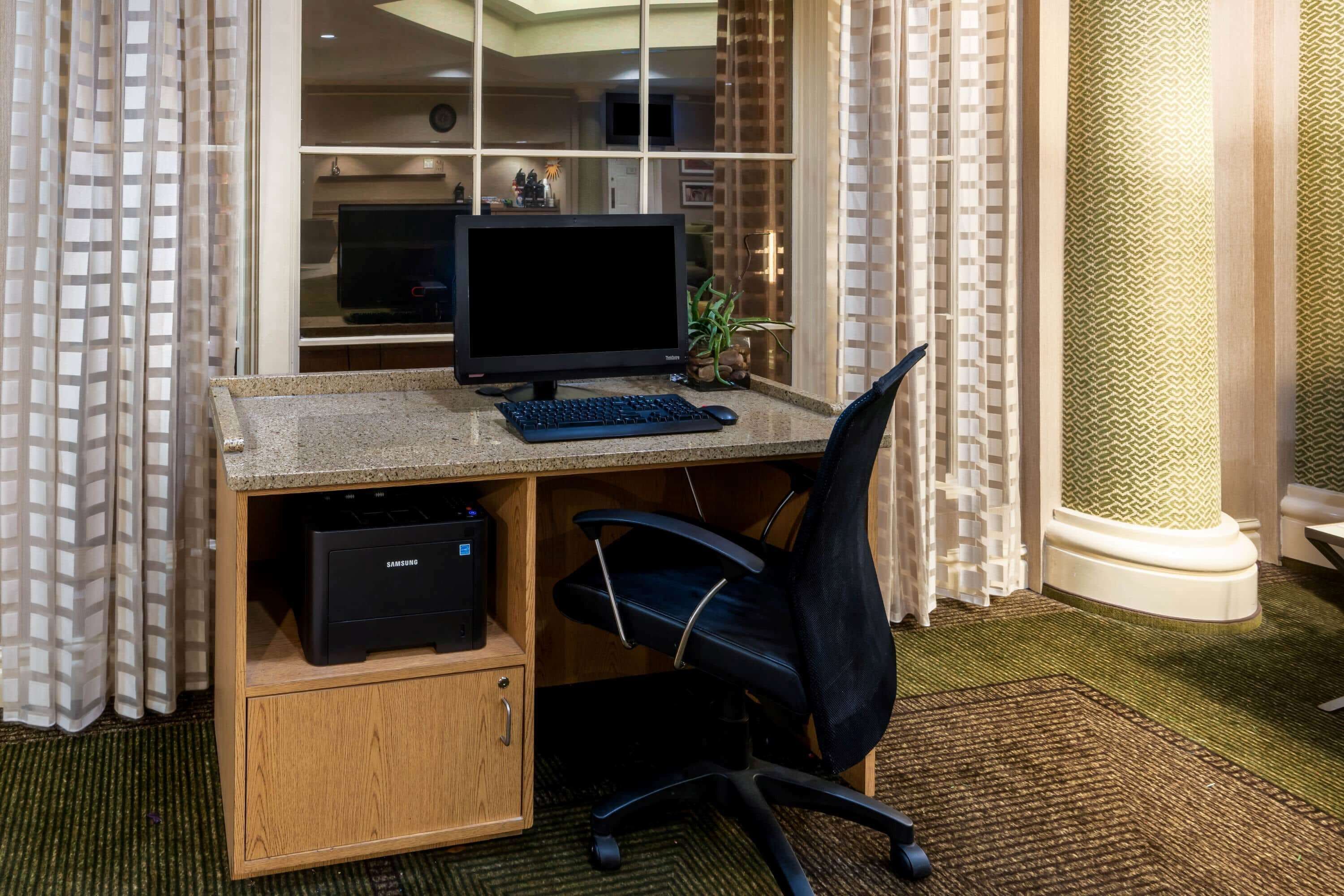 A home office setup with a desktop computer, a black office chair, a printer, and a bookshelf.