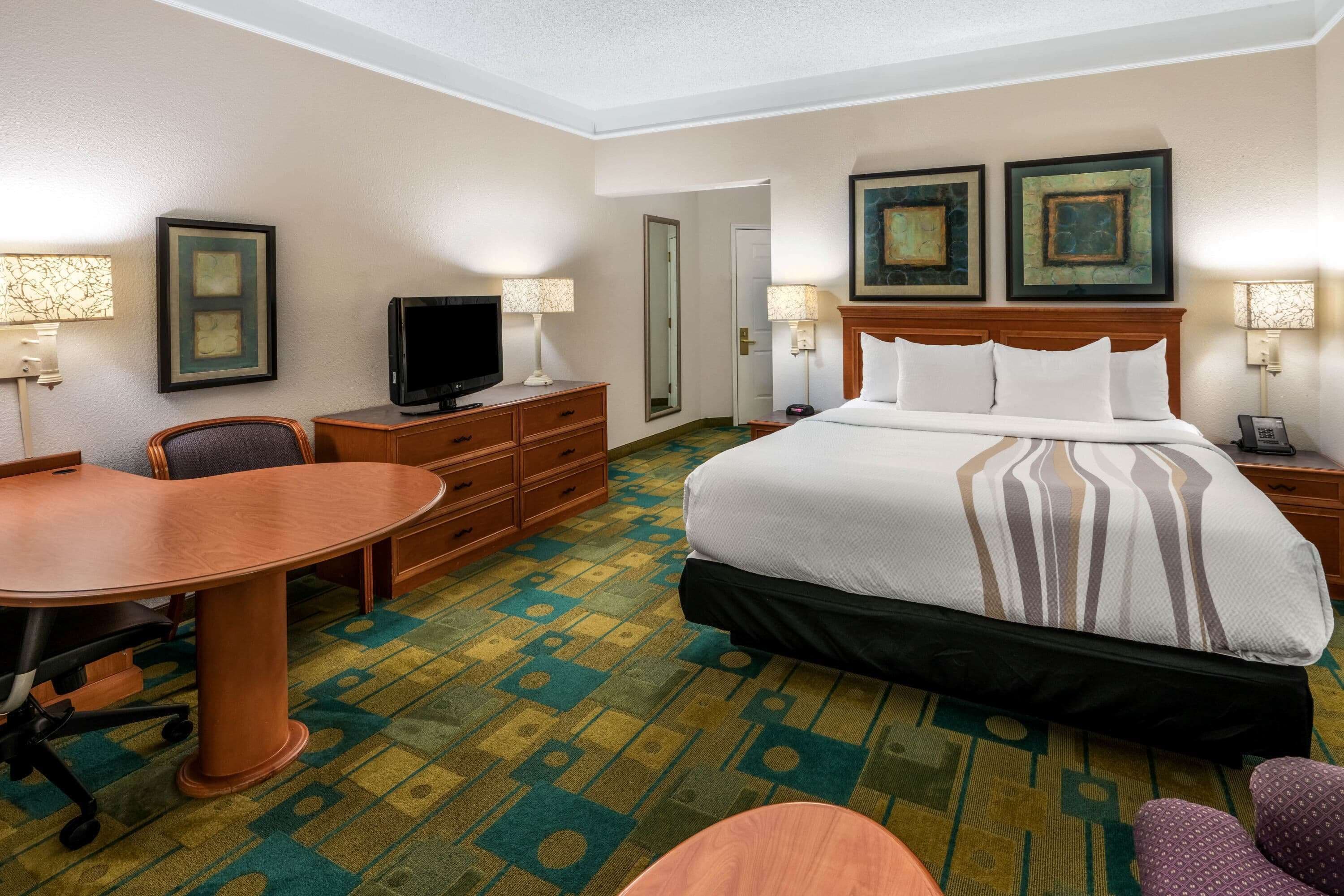 A hotel room with a bed, a desk, a chair, a television, and a carpet with a geometric pattern.