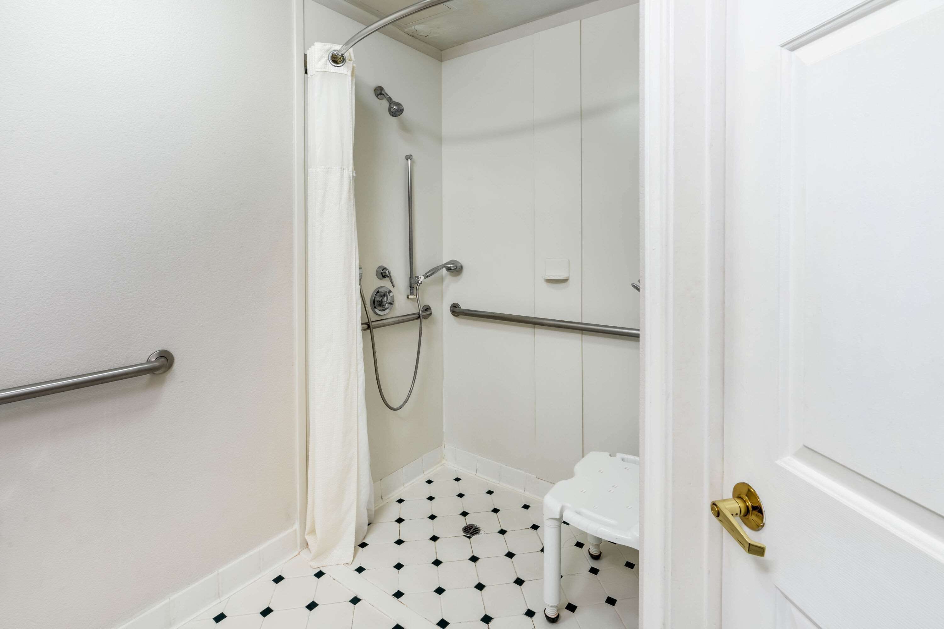 A small, white bathroom with a shower area, a white sink, and a door with a brass handle.