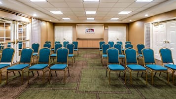 Un salón de conferencias con filas de sillas azules, suelo alfombrado, puertas blancas y un cuadro enmarcado en la pared.