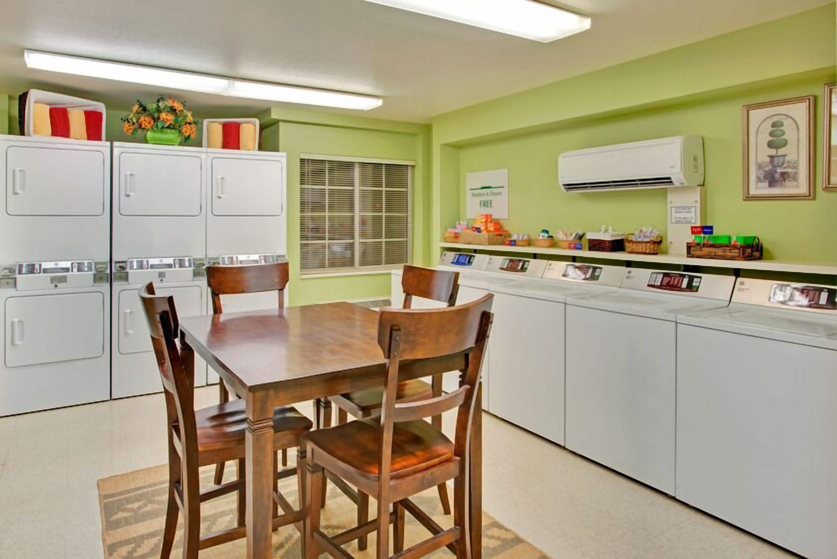 A compact kitchen with a dining table and chairs, white cabinets, a window with blinds, and a wall-mounted air conditioner.