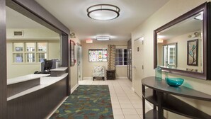 A hotel reception area with a reception desk, a waiting area with a chair, a bulletin board, and a hallway with a patterned carpet.