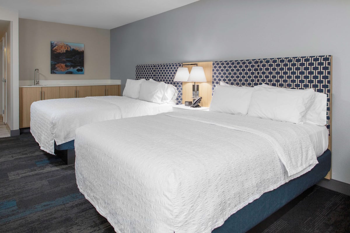 A hotel room with two beds, a nightstand with a lamp, a painting on the wall, and a bathroom with a sink and mirror.