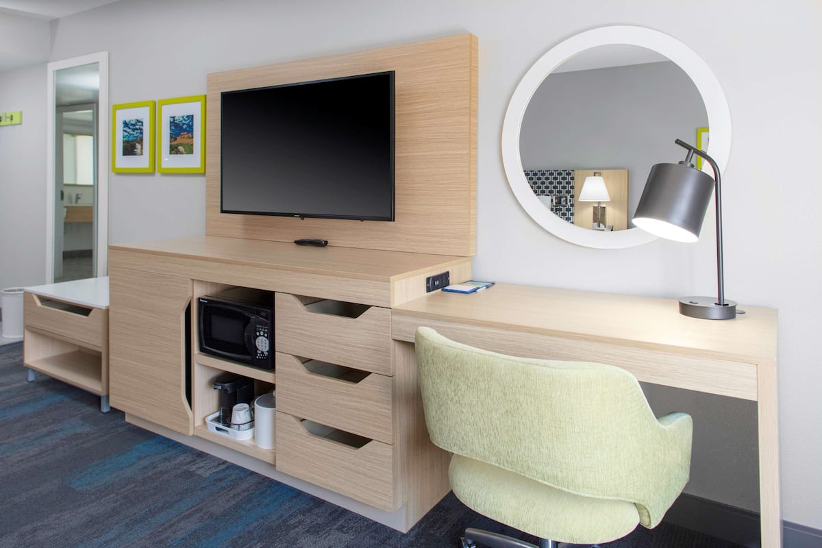 A modern hotel room with a wooden desk, a flat-screen TV, a round mirror, and a green chair.