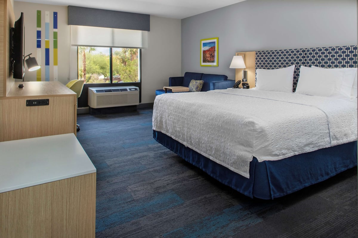 A hotel room with a large bed, a desk, a chair, a TV, and a window with a view of trees.