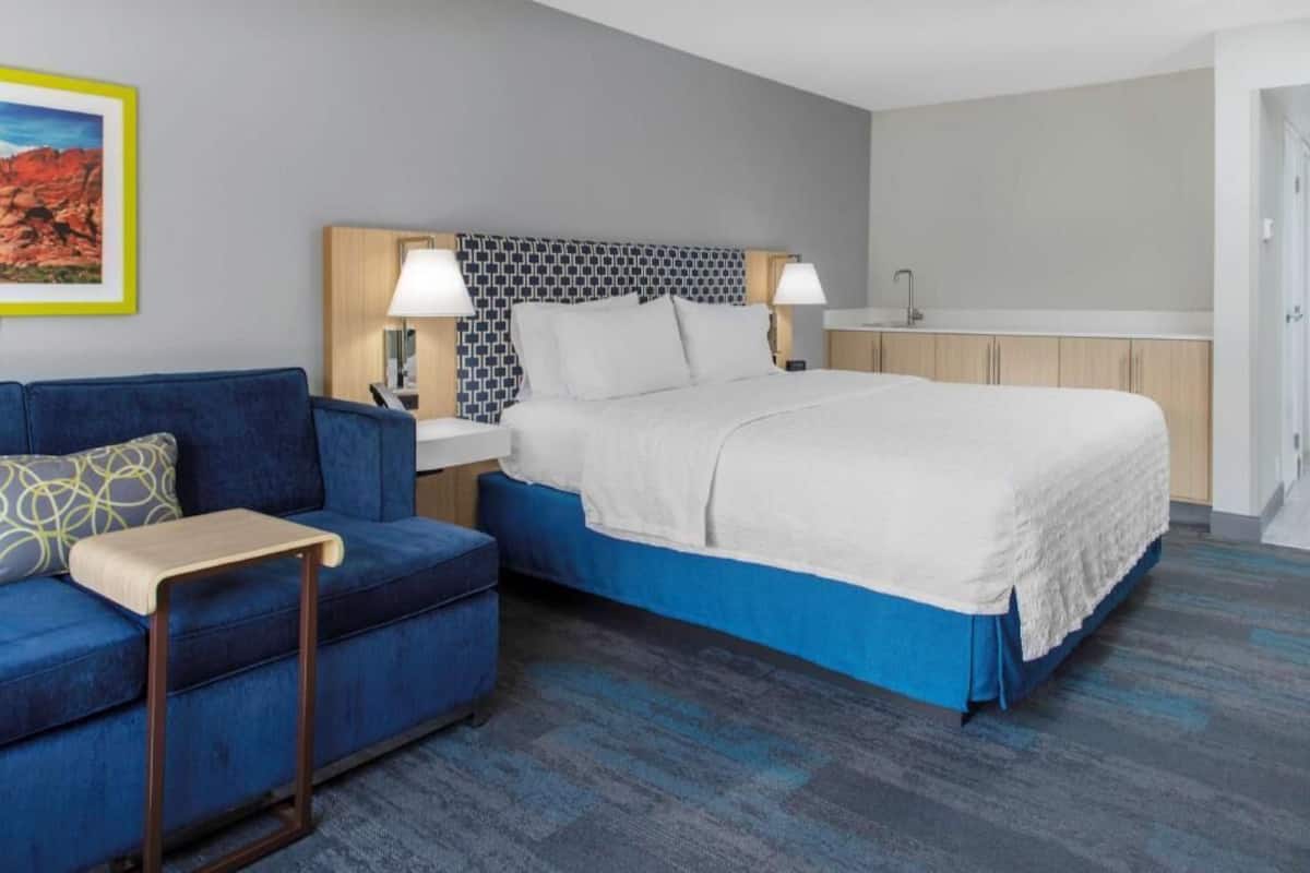 A modern hotel room with a bed, a sofa, a small table, a nightstand, and a bathroom.