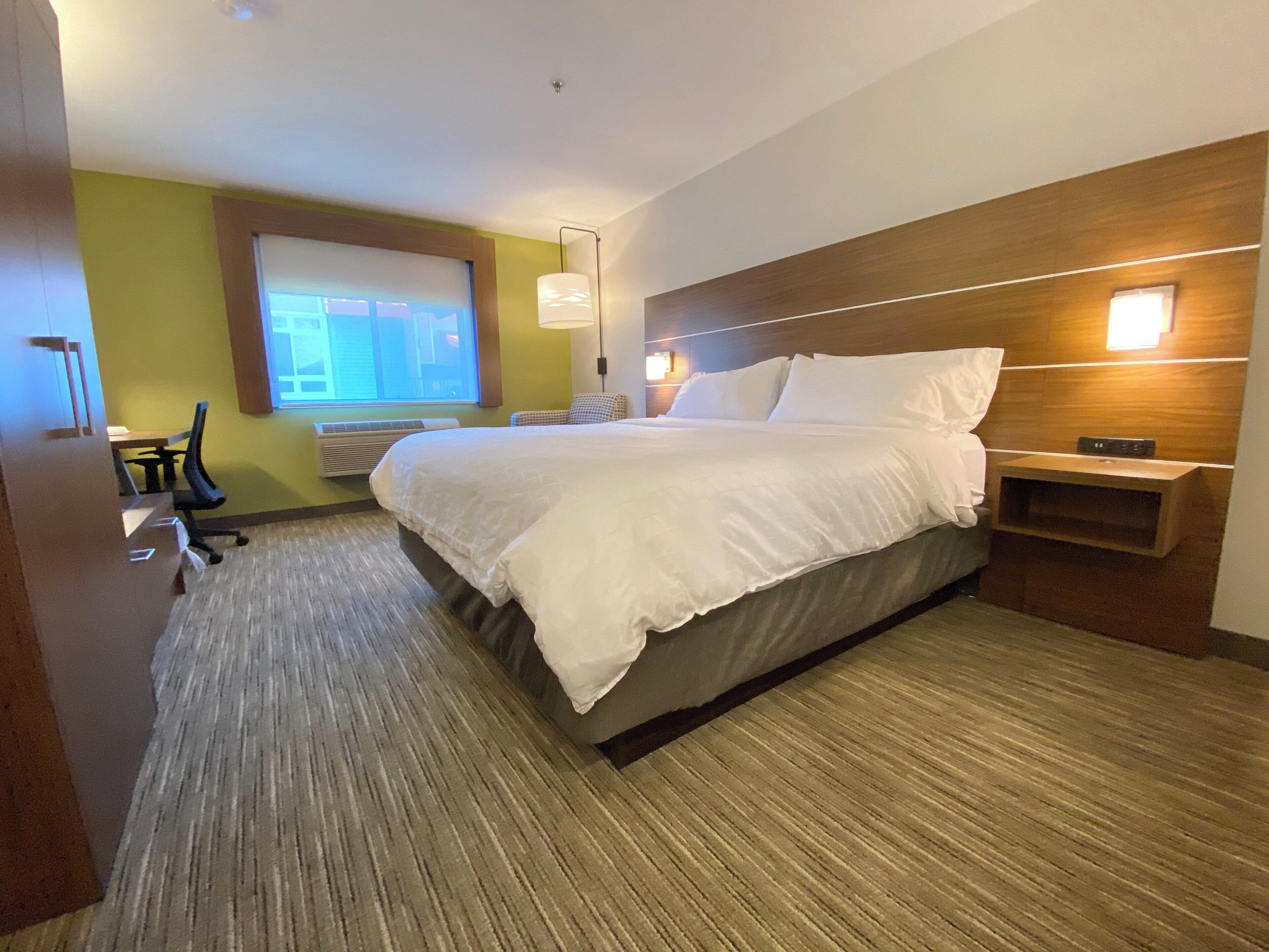 Habitación de hotel con una cama grande, un escritorio, una silla, una ventana y una mesita de noche fijada a la pared.