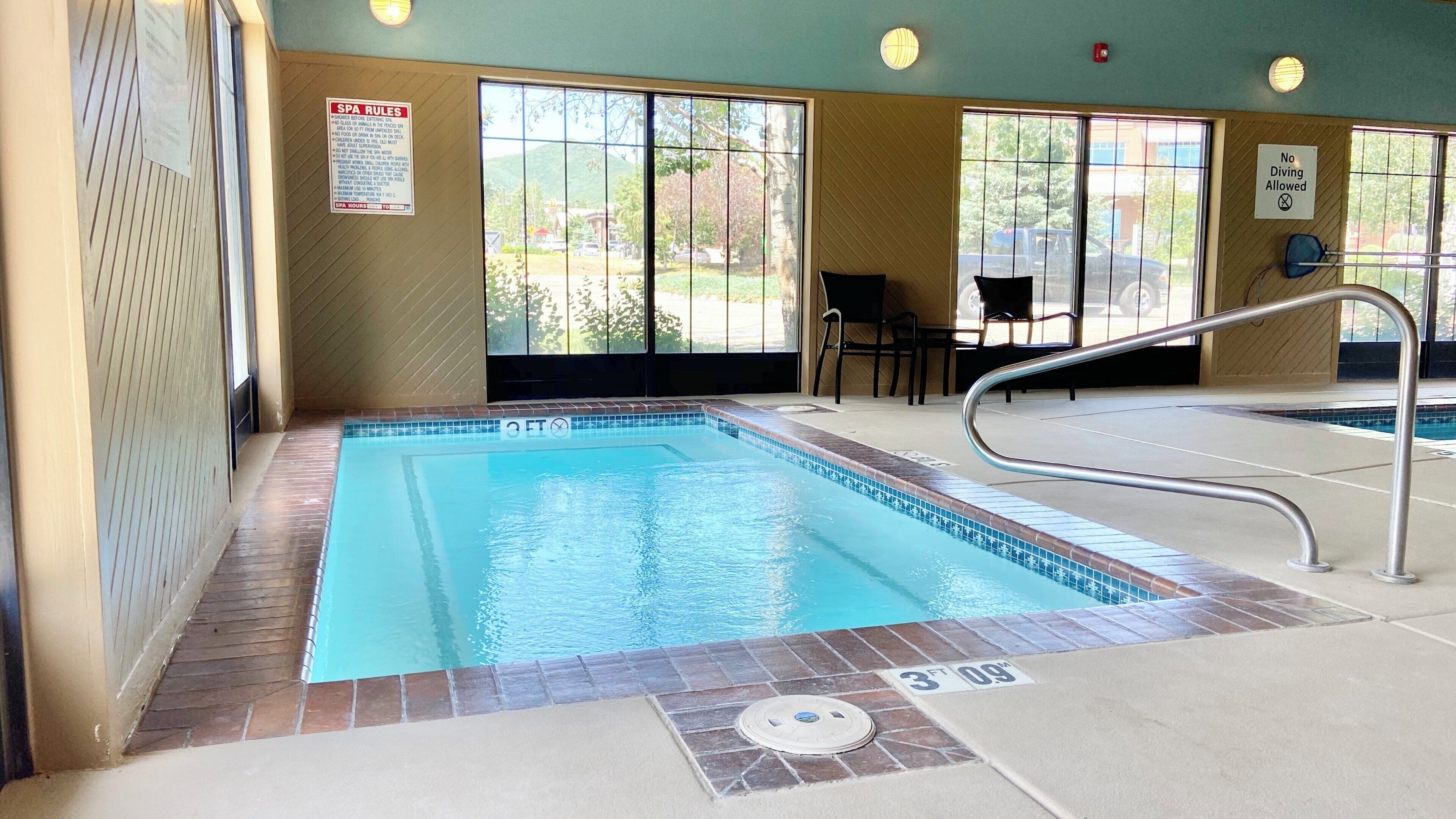A swimming pool with a metal ladder, a sign indicating no diving allowed, and a view of the outside through large windows.