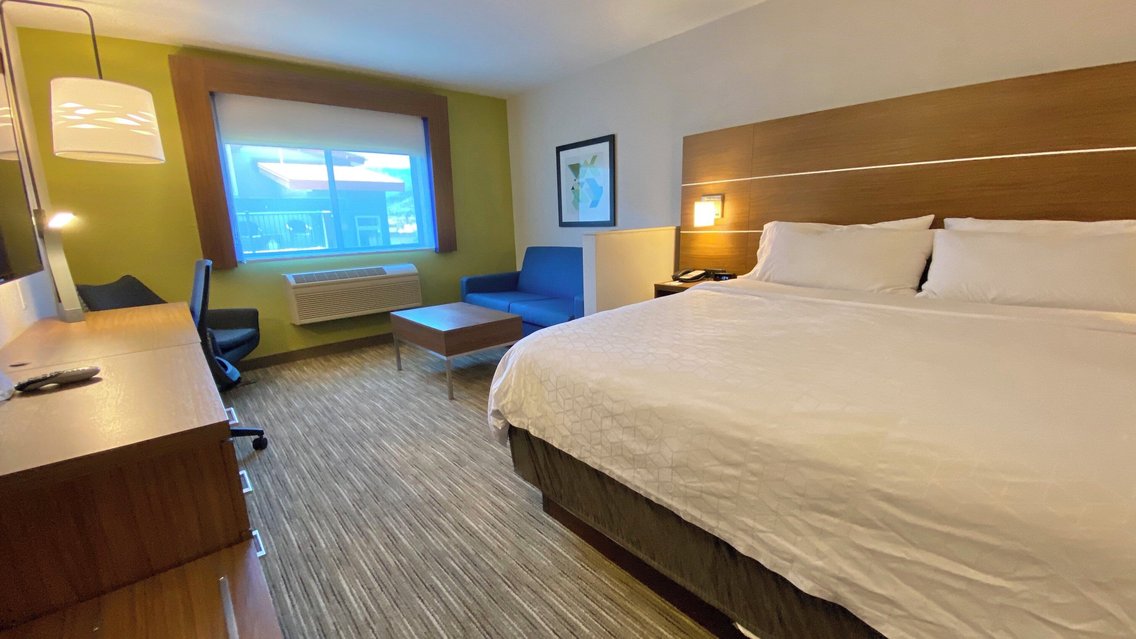 Une chambre d’hôtel avec un grand lit, un bureau avec une chaise, un canapé bleu, une fenêtre donnant sur un beau paysage et un tableau encadré au mur.