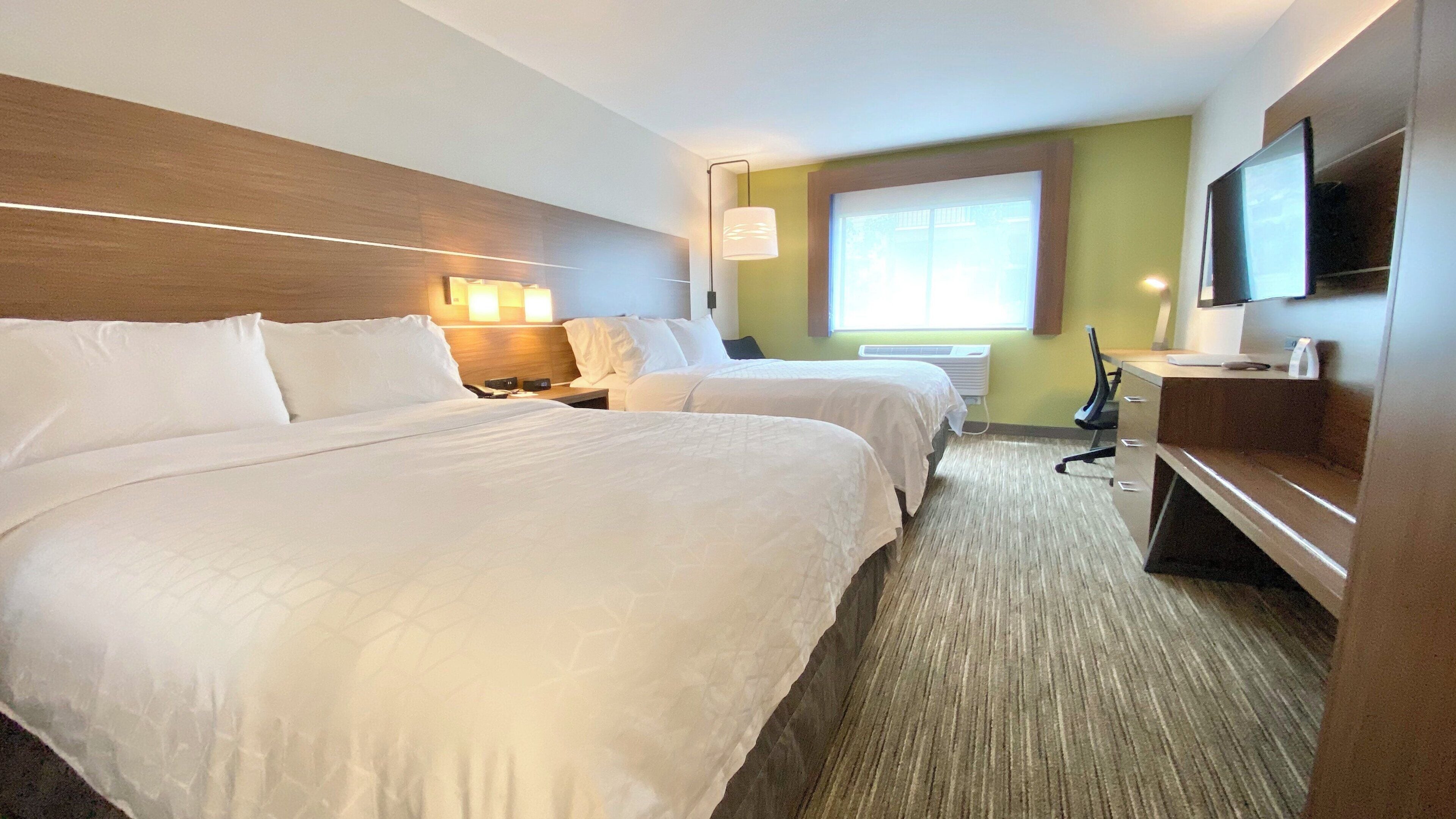 Une chambre d’hôtel avec deux lits, un bureau, une chaise, une télévision et une fenêtre avec des stores.