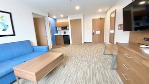 Habitación de hotel moderna con sofá azul, banco de madera, kitchenette, baño y televisor.