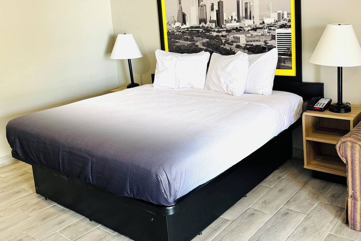 A hotel room with a large bed, two bedside lamps, a nightstand with a phone, and a framed cityscape picture on the wall.