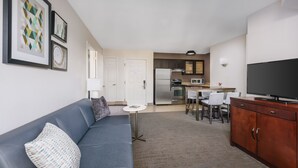 A modern living room with a grey sofa, a coffee table, and a TV. The kitchen area is visible in the background with a refrigerator, microwave, and dining table. - Residence Inn by Marriott Gaithersburg Washingtonian Center (Gaithersburg)
