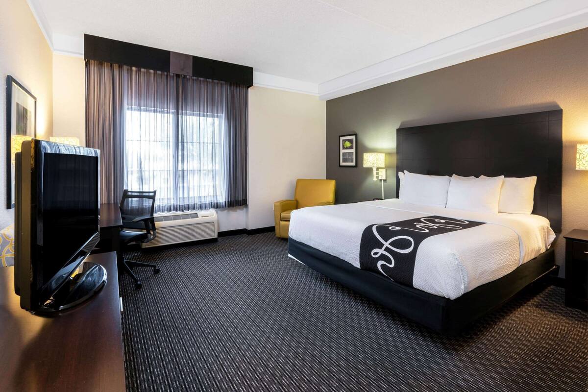 Une chambre d’hôtel avec un grand lit, un bureau avec un ordinateur, une chaise, une télévision et une fenêtre avec des rideaux.