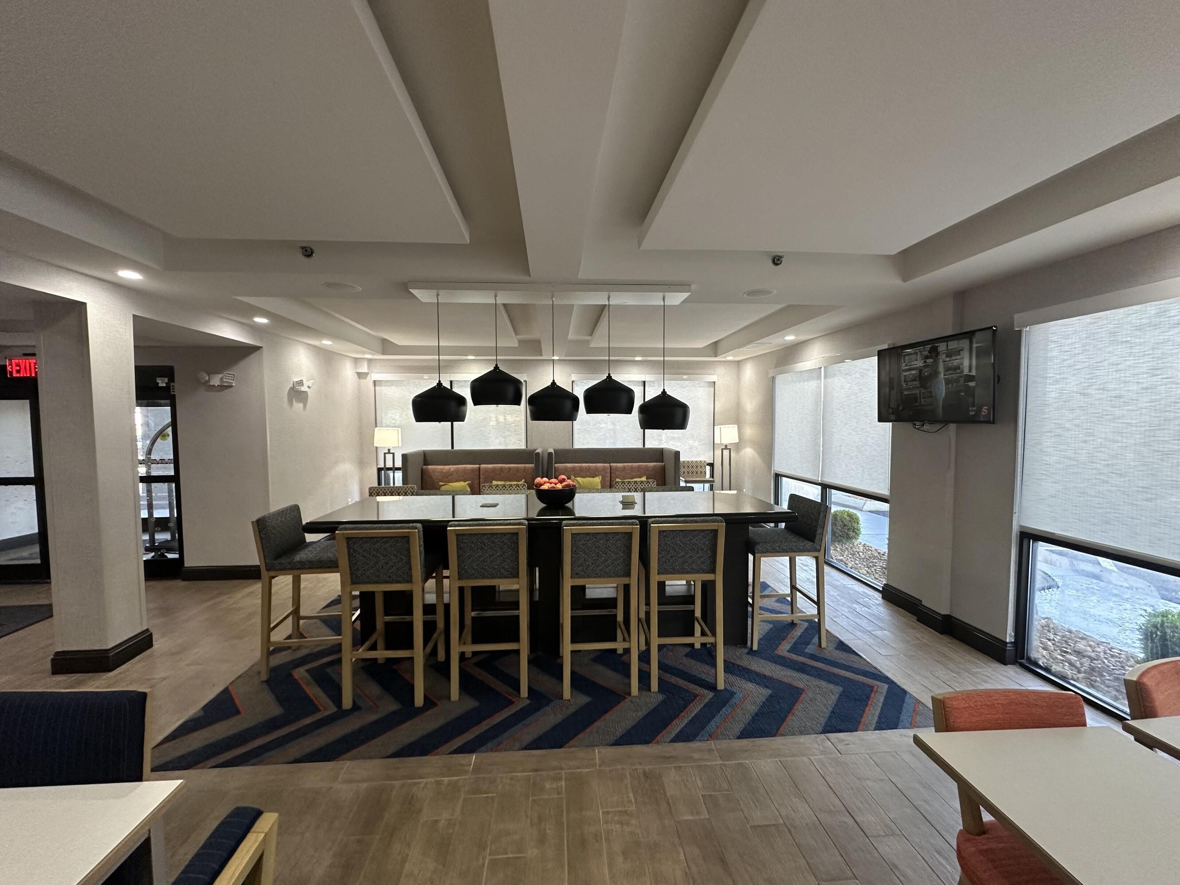 A modern hotel room with a dining area featuring a large table and chairs, a sofa, and a television mounted on the wall.
